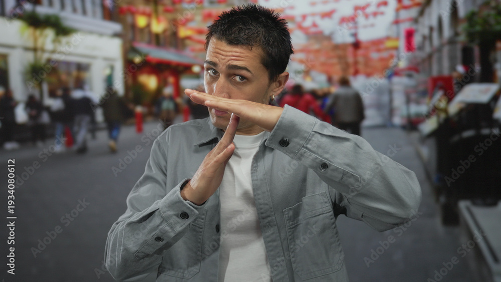 Fototapeta premium Young man making time-out gesture on bustling urban street with festive decorations and diverse crowd walking by