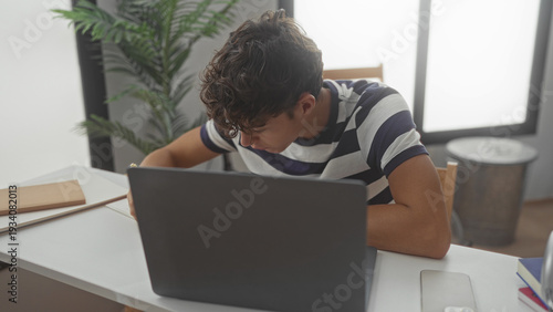 Billede på lærred Teen student leans over laptop writing with pencil at studio desk, head bent and arm resting on table; study concentration