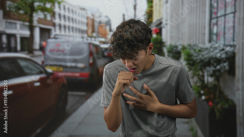 Photos Teenage boy in grey t shirt clutching throat and coughing on a busy city street beside parked cars and a delivery van; distress urgency