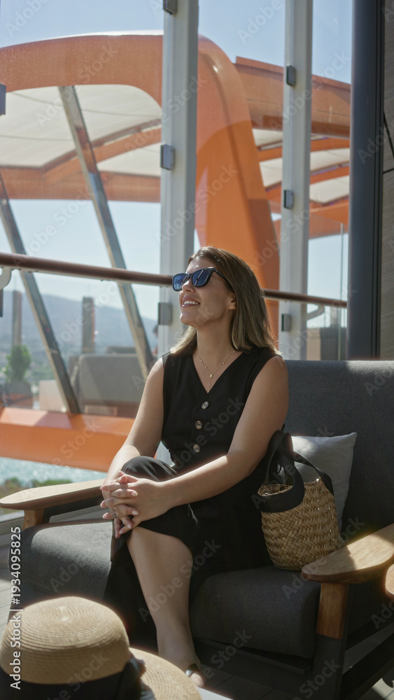 Obraz premium Woman holding sunglasses on cruise terrace lounge chair with straw bag and hat beside her while she sits and reaches toward hat; travel leisure calm.