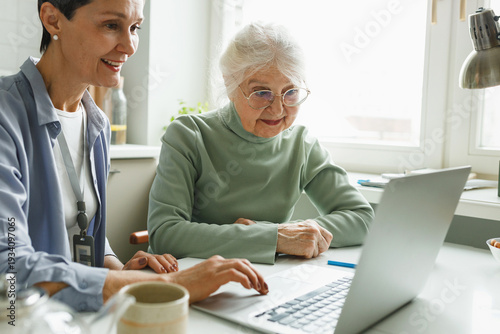 Two people using laptop, elderly lonely female in glasses learning from her caregiver how to use online applications, web-pages, social media and messengers both sitting in kitchen with window