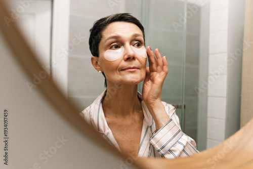 Fotografija Closeup of mirror reflection of mature good-looking female with short hair applying eye patches in evening to moisturize skin and get rid of wrinkles, patting face