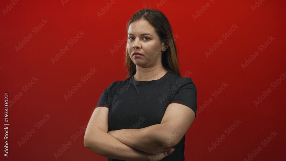 Obraz premium Woman young caucasian with arms crossed and a stern expression, wearing black shirt and direct gaze in red studio; annoyance.