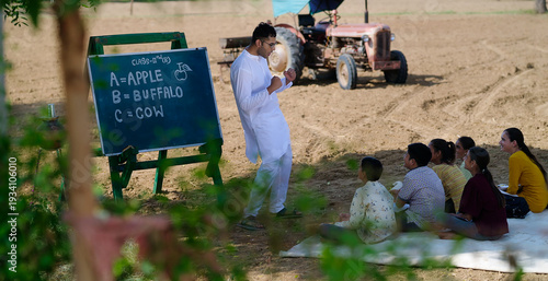 Indian young adult male man do dance teach villager girl boy standing under tree rural ground area. Happy child group sitting on mat look tutor learn enjoy new day have fun joy open class field place