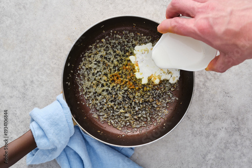 Cooking onions and spices in a pan while adding cream in a kitchen setting during daytime hours