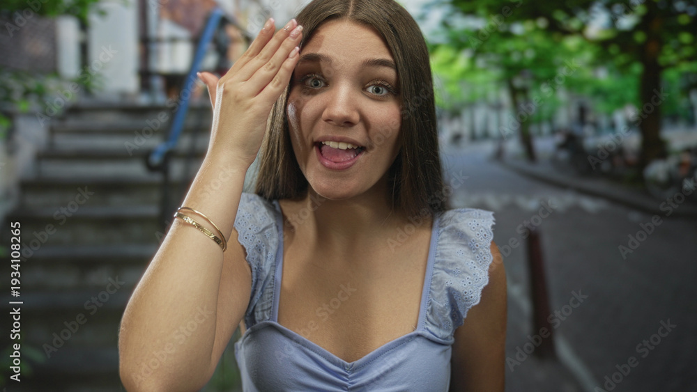 Fototapeta premium Caucasian teenage girl in blue top covers eyes with hand while smiling on sunlit urban street; shyness mystery.