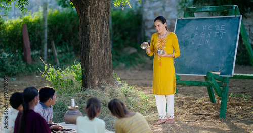Indian happy female teach boy group do study sitting on mat rural area young girl show tiffin box in hand talk speak gossip enjoy new day desi life spend day break time outdoor open class air field