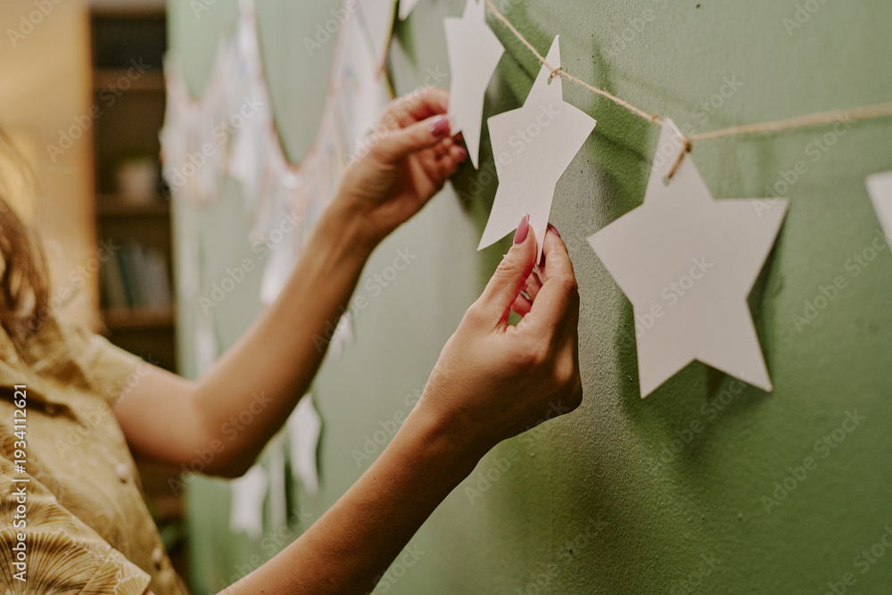 Obraz premium Caucasian young adult woman decorating wall by hanging paper star garland, hands carefully arranging decorations on string, preparing festive display against green background