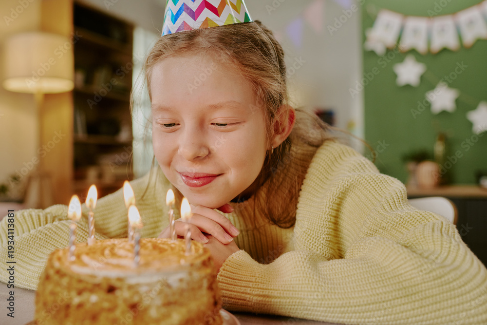 Fototapeta premium Caucasian girl child wearing party hat smiling and gazing at birthday cake with lit candles, resting chin on hands, celebrating birthday indoors with festive decorations in background