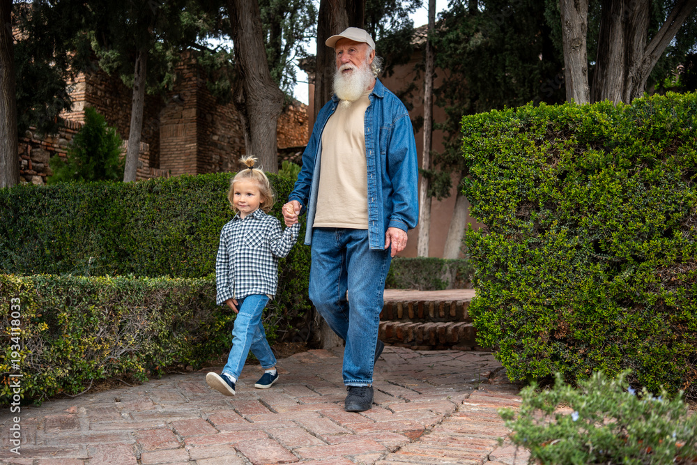 Fototapeta premium A grandfather and his grandchild walking hand in hand through a garden with neatly trimmed hedges and trees in the background