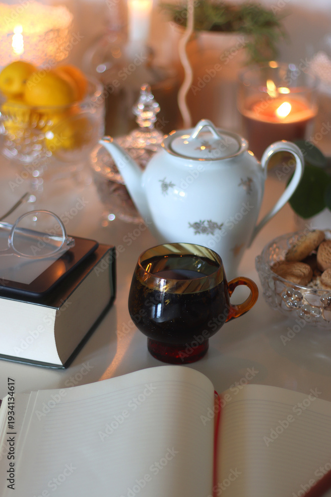 Obraz premium Cup of tea or coffee, teapot, book, notebook, e-reader, glasses, cookies, nuts, lemons, house plants and lit candles on the table. Hygge at home. Selective focus.