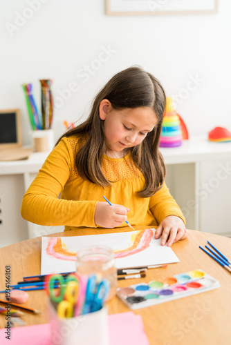 Little girl using watercolors to paint a rainbow wearing a yellow shirt in a bright and colorful home