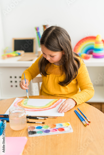 Little girl using watercolors to paint a rainbow wearing a yellow shirt in a bright and colorful home