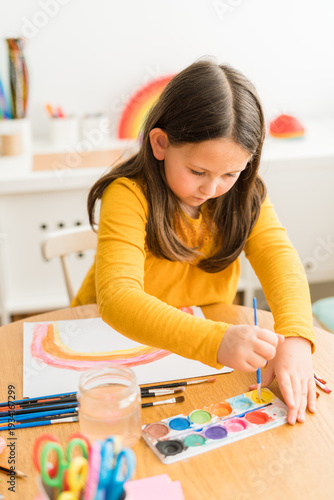 Little girl using watercolors to paint a rainbow wearing a yellow shirt in a bright and colorful home