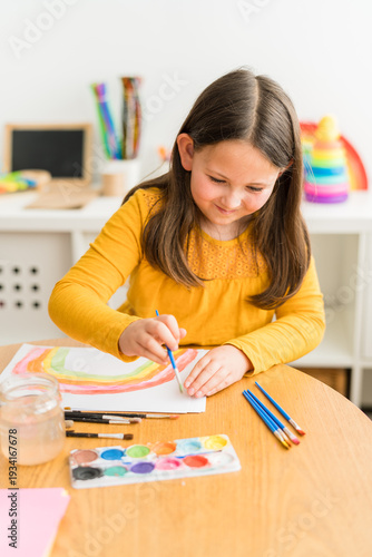 Little girl using watercolors to paint a rainbow wearing a yellow shirt in a bright and colorful home