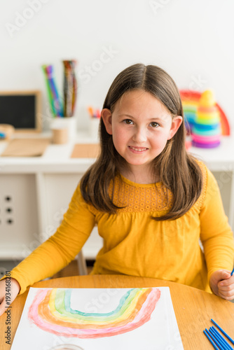 Little girl smiling and looking at camer while using watercolors to paint a rainbow wearing a yellow shirt in a bright and colorful home