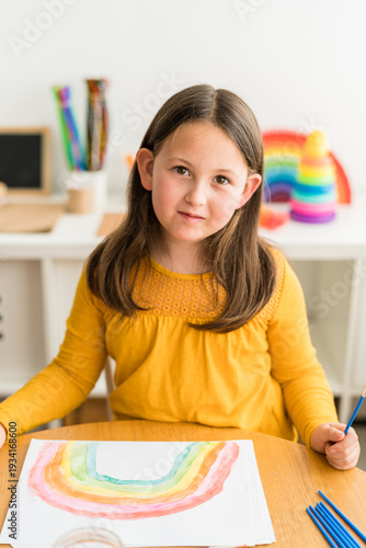 Little girl smiling and looking at camer while using watercolors to paint a rainbow wearing a yellow shirt in a bright and colorful home