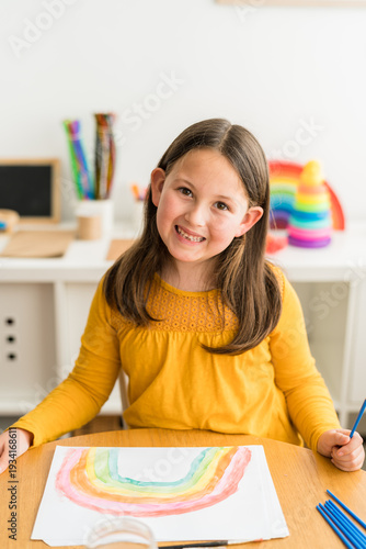 Little girl smiling and looking at camer while using watercolors to paint a rainbow wearing a yellow shirt in a bright and colorful home