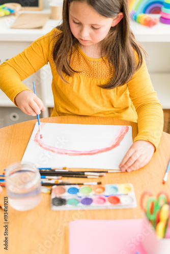 Little girl using watercolors to paint a rainbow wearing a yellow shirt in a bright and colorful home