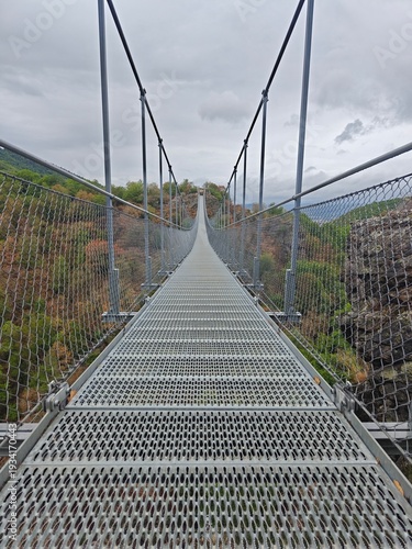 Long modern metal suspension bridge with a unique perforated walkway extends across a scenic valley.