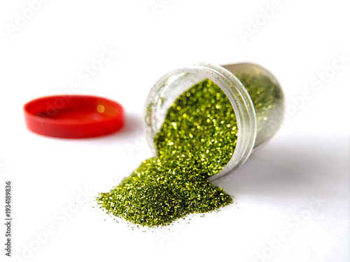 Green glitter spills from a small clear plastic container, creating a shimmering pile on a white surface.