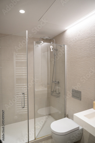 Modern bathroom with frameless glass shower, rain and handheld heads, floating toilet and integrated white sink countertop against textured beige tiles and recessed lighting