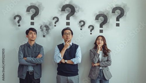複数の疑問符が浮かぶ壁の前で今後の方針に悩む東アジア人ビジネスチーム三人