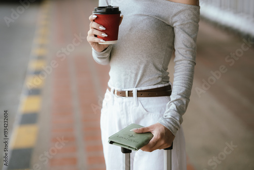 Traveler holding coffee cup and smartphone with yellow suitcase at airport curbside, waiting for transportation before departure.