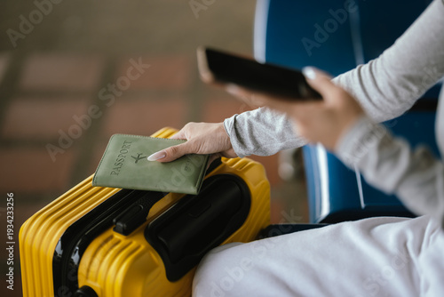 Close-up of traveler holding passport and smartphone on yellow suitcase while waiting at airport terminal, preparing for flight.