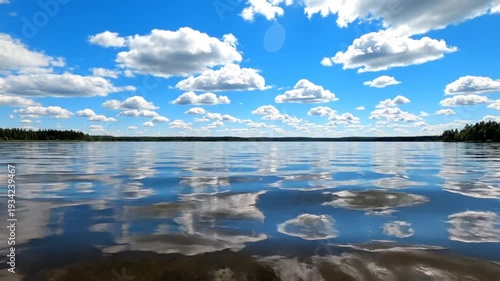 A serene lake reflecting a bright blue sky filled with fluffy white clouds, with a forest in background
