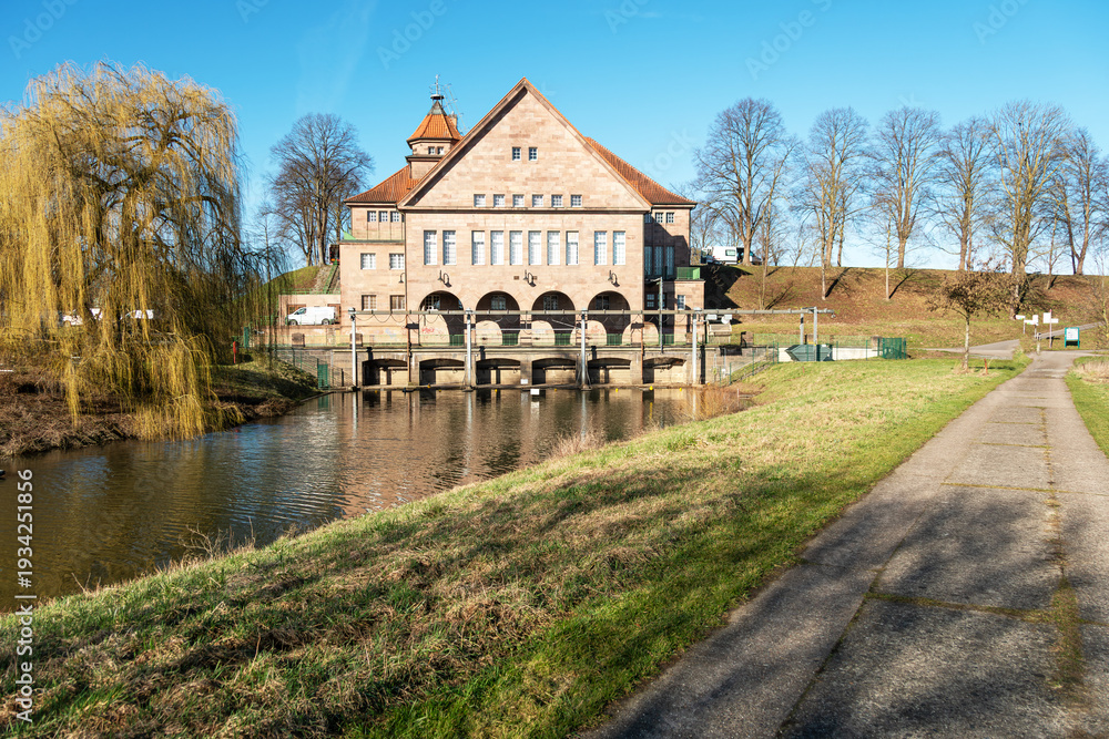 Obraz premium The massive pumping station rises beside the calm canal while fresh green tones and a willow tree suggest early spring. Architecture and hydraulic engineering in clear perspective.