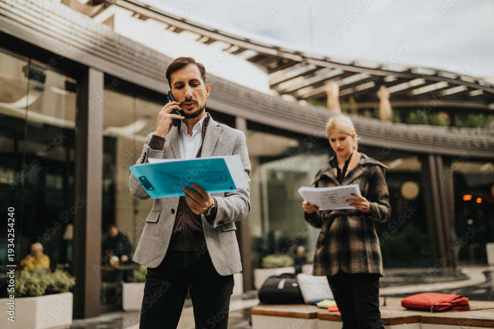 Fototapeta premium Two professionals outside a modern building review documents and speak on the phone, conveying collaboration and business activity.