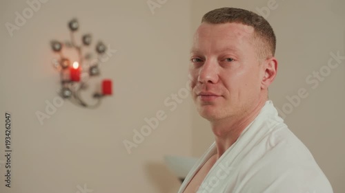 white man in robe by sconce, spa client smiles between profile and closeup during professional microcurrent facial, warm candlelight on beige wall, modern lamp and mirror in background, relaxed