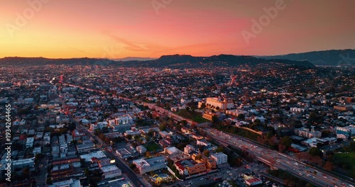 Approaching multi-lane highway with hectic traffic crossing the vast city scenery. Panorama of Los Angeles, California, USA against pink sky. Mountain silhouettes along the horizon. Aerial view.