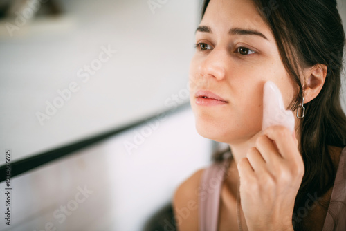 A young woman makes a facial massage with a gouache scraper at the mirror at home.