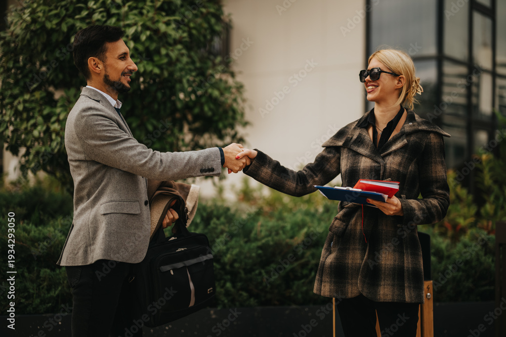 Obraz premium Two professionals greet with a handshake in an outdoor setting, smiling, with folders and a briefcase nearby.