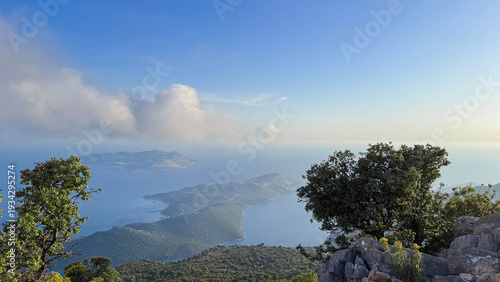 A summer panoramic view from the mountains, the Mediterranean coast, the town of Kas, Turkiye. Green hills, a bay with blue water, and a cloudy sky.
