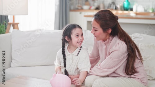 Joyful Asian mother teaches her young daughter how to save money by putting a coin into a pink piggy bank together in a bright modern living room.