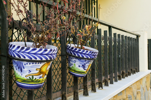 Two decorative ceramic pots with withered plants hanging on a metal railing outdoors