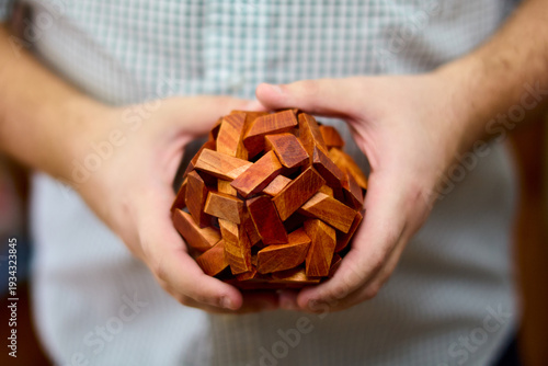 Quadro em tela Hands holding a wooden interlocking puzzle sphere made of reddish-brown rectangular blocks, checkered shirt background