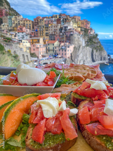 Mixed bruschetta platter with pesto, tomato, mozzarella, burrata, melon and focaccia on a terrace with colorful houses of Manarola blurred in the background, Cinque Terre, Liguria, Italy
