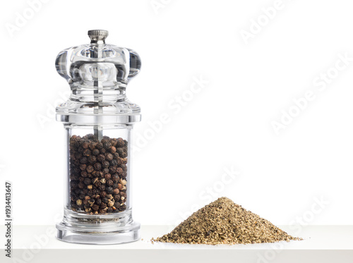 Glass pepper grinder filled with black peppercorns and pepper ground on white table.
