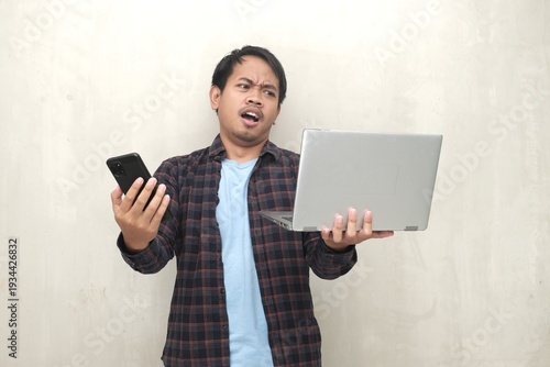 Southeast Asian man holding laptop and smartphone, looking confused and complaining about tech problems, isolated on plain wall background studio.