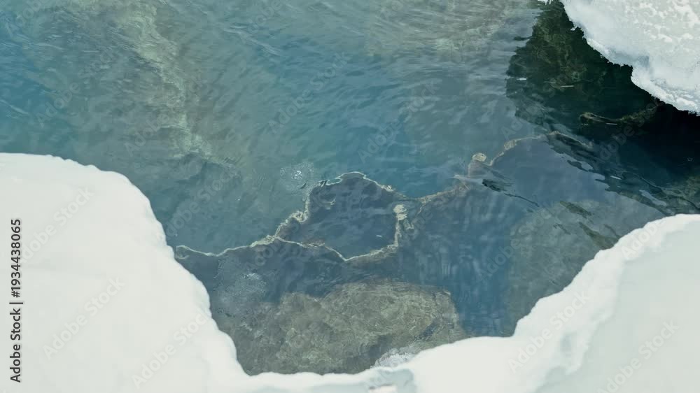 Clear water shows the rocky bottom of a frozen lake with ice around the ...