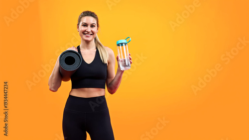 Smiling fitness woman holding yoga mat and water bottle on orange background