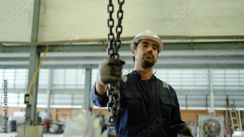 Professional male industrial technician in safety gear operates a heavy duty chain hoist and crane remote control inside a large manufacturing factory or modern warehouse facility.
