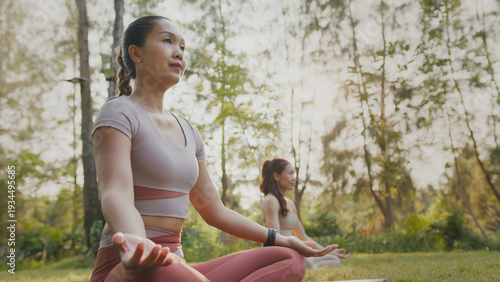 Two women meditate in outdoor park at sunrise with hands pressed together, calm focus, soft light and yoga mat on grass