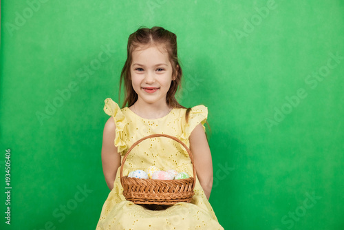 Little girl smiles while holding a wicker basket with pastel-colored Easter eggs, set against a vibrant green backdrop