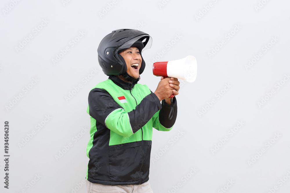Fototapeta premium Asian online motorbike taxi driver man speaking facing sideways using megaphone giving announcement, news or advertising promotion, isolated on grey background.