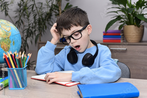 Exhausted child doing homework at home, holding head with hand, surrounded by books, pencils and globe. Education, school stress, concentration problem and modern learning concept.